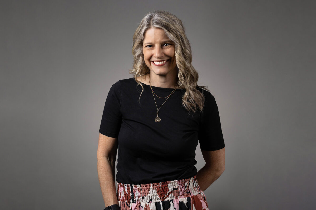 Woman with long blonde hair wearing a black shirt and patterned skirt, smiling at the camera against a plain background, captured by Denver Headshot Co.