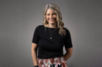 Woman with long blonde hair wearing a black shirt and patterned skirt, smiling at the camera against a plain background, captured by Denver Headshot Co.