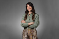 A person with long, dark hair wearing a green shirt and patterned pants stands against a gray background, smiling with arms crossed—a perfect capture from Denver Headshot Co.