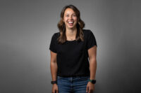 A person stands against a grey background, smiling while wearing a black shirt, blue jeans, and two wristbands. This portrait perfectly captures the professionalism Denver Headshot Co is known for.