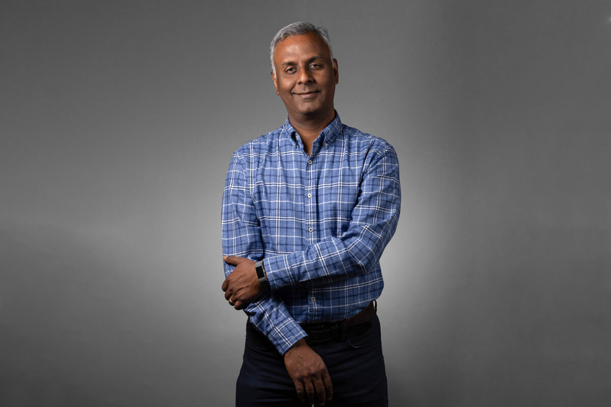 A man wearing a blue plaid shirt stands against a plain grey background with his arms crossed, smiling lightly—a perfect shot by Denver Headshot Co.