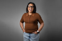 A woman in a brown short-sleeve shirt and jeans, wearing glasses, smiles with her hands in her pockets against a plain grey background. This candid shot by Denver Headshot Co perfectly captures her approachable and confident demeanor.