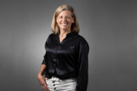 A woman with shoulder-length blonde hair wearing a black blouse and white skirt, captured expertly by Denver Headshot Co, stands against a plain gray background, smiling at the camera.