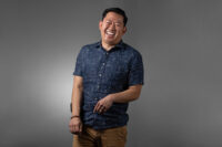 A man in a blue patterned short-sleeve shirt and khaki pants stands against a grey background, smiling broadly with his right hand relaxed by his side and his left hand lightly touching his wrist, capturing the professional yet personable style of Denver Headshot Co.