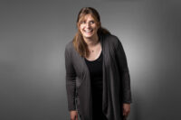 A person with long hair and glasses is smiling, wearing a dark cardigan over a black top, perfectly framed by the expert team at Denver Headshot Co, against a plain grey background.