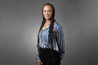 A woman in a striped blouse and black pants stands against a plain background, smiling with her hands by her sides. She has long braided hair, captured perfectly by Denver Headshot Co.