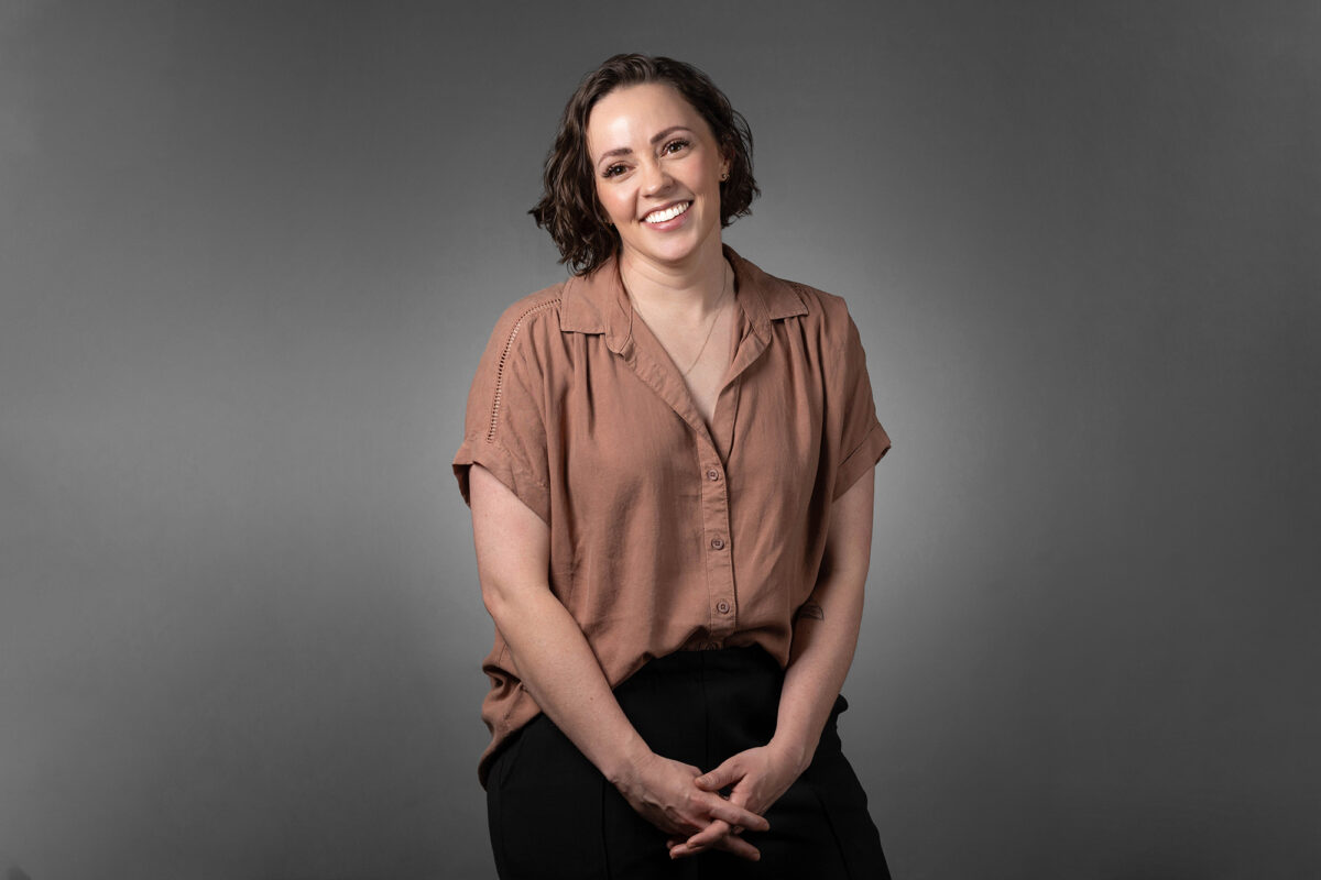 A person with short hair and a beige button-up shirt smiles while sitting against a gray background in this professional Denver Headshot Co portrait.