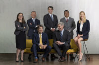 A group of seven professionally-dressed individuals pose for a formal photo by Denver Headshot Co. Three are seated on a yellow couch, and four stand behind them against a plain background.