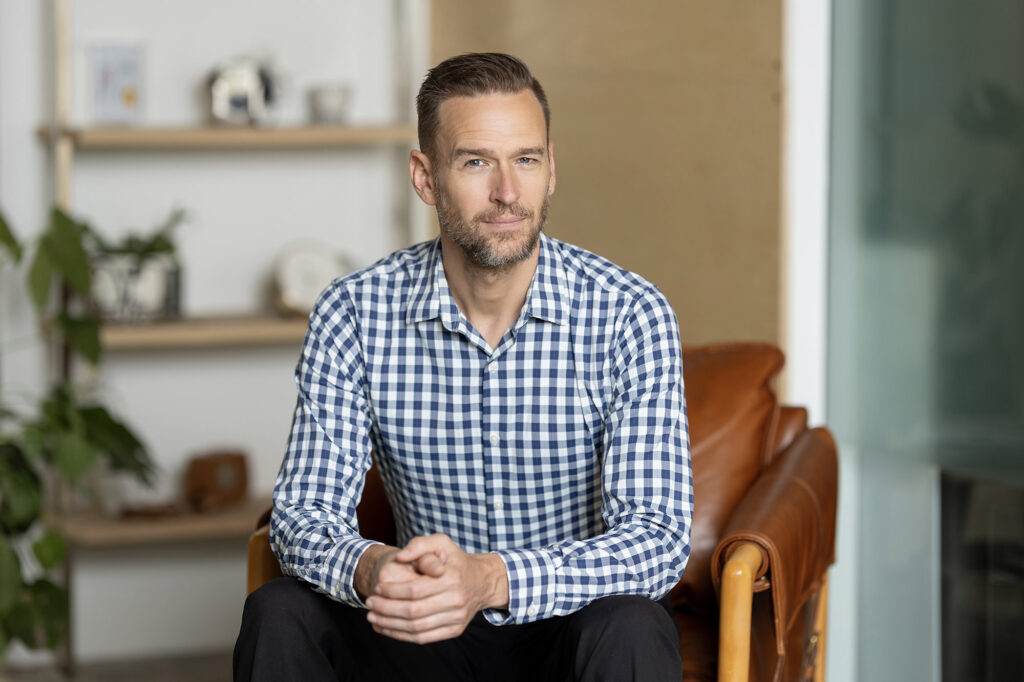 A man with short hair and a beard is wearing a checkered shirt, seated on a brown leather chair. Shelves with decorative items and a plant adorn the background, creating the perfect setting for a Denver Headshot Co session.