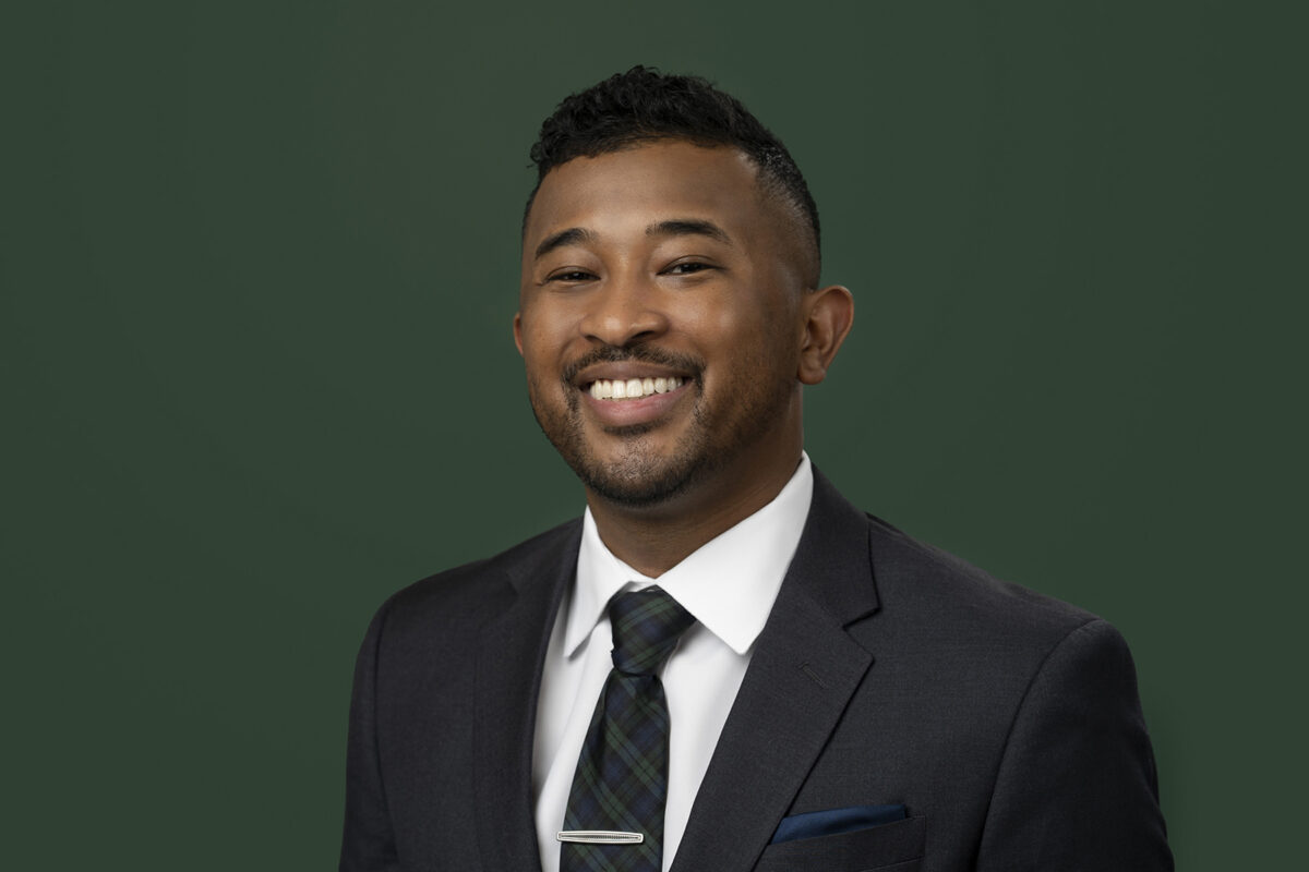 A man wearing a dark suit, white shirt, and patterned tie, smiling confidently against a plain dark green background — showcasing the professional touch of Denver Headshot Co.