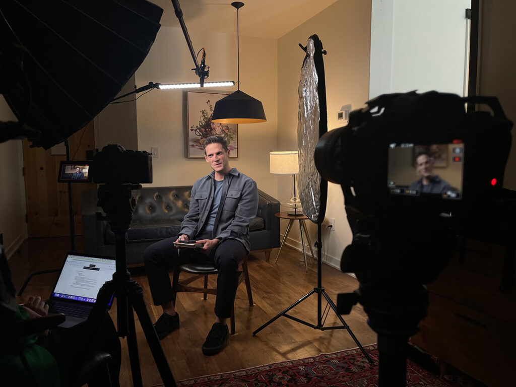 A man sits on a chair in a well-lit room set up for video recording, with cameras, lights, and a laptop visible in the foreground. The setup hints at the professionalism of Denver Headshot Co.