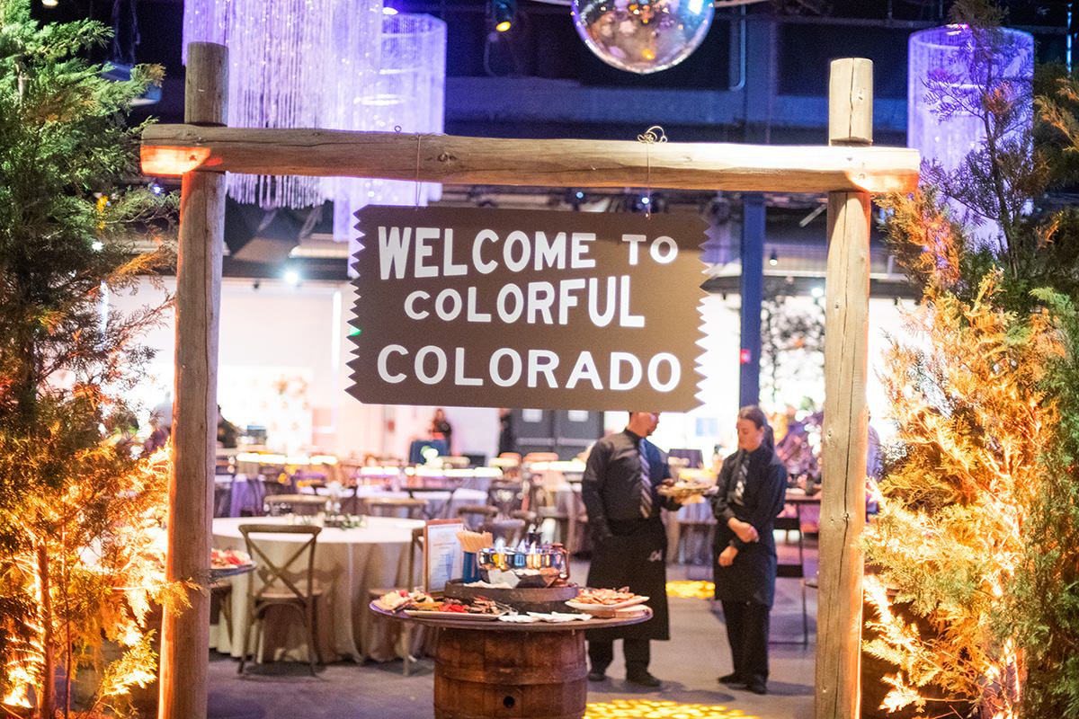 A wooden sign reads "Welcome to Colorful Colorado" at an indoor event decorated with trees, lights, and a table of food. Two people are standing in the background, capturing memories courtesy of Denver Headshot Co.