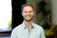 A smiling man with short hair and a beard wears a patterned shirt and stands indoors, captured in a sharp image by Denver Headshot Co., with a beautifully blurred background.