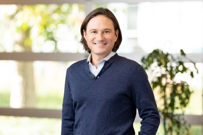 A person with shoulder-length hair, wearing a blue sweater and striped shirt, stands indoors with a smile. There's a window and a plant in the background, capturing an inviting ambiance perfect for Denver Headshot Co.