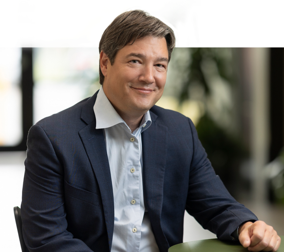 A man in a dark blazer and light blue shirt is seated, smiling, and looking towards the camera. Photographed by Denver Headshot Co, there is a blurred background with greenery.