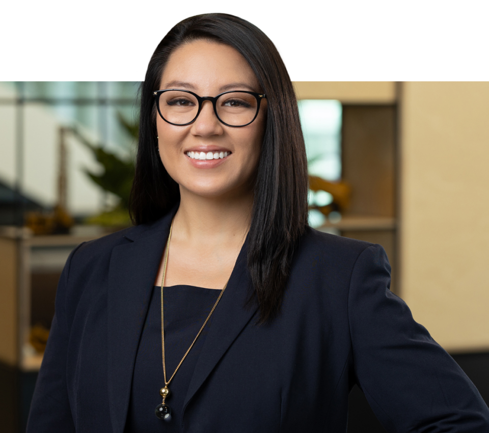 A woman with long dark hair and glasses, wearing a navy blue blazer and a gold necklace, stands smiling in an office environment—captured perfectly by Denver Headshot Co.