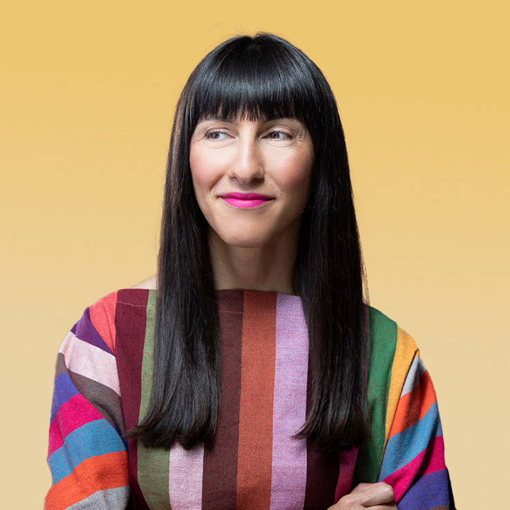 A woman with long black hair and bangs, wearing a multicolored striped top, stands against a yellow background with her arms crossed and looking slightly to the side. Captured by Denver Headshot Co.