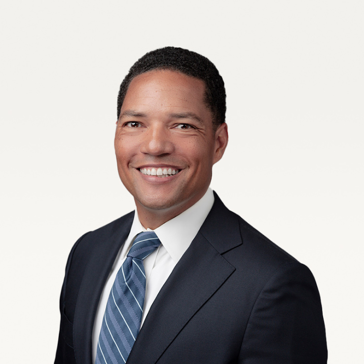 Portrait by Denver Headshot Co of a smiling man wearing a dark suit, white shirt, and a striped blue tie with a plain light background.