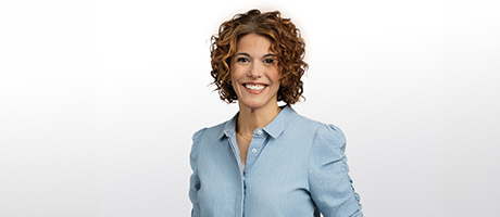 A woman with curly hair, wearing a light blue blouse, smiles at the camera against a plain white background.