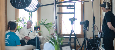 A behind-the-scenes view of an interview setup with lighting, cameras, microphones, and a technician overseeing the recording. Two people are seated and engaged in conversation.