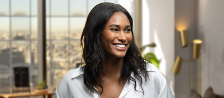 A woman with long dark hair and a white shirt smiling in a well-lit modern office with large windows and cityscape views in the background.