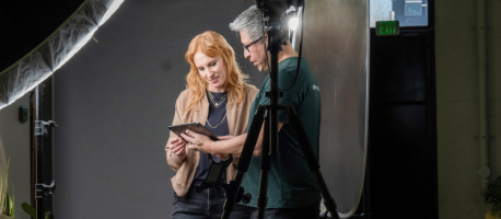 Two people stand in a photo studio, looking at a tablet. One person with long red hair holds the tablet, while the other, with short gray hair, looks on. Studio equipment is visible around them.