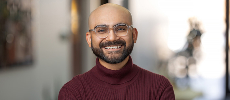 A person with a beard and glasses is smiling while wearing a maroon sweater. They are standing indoors with a soft-focus background.