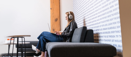 A person sitting on a grey couch wearing headphones and using a laptop.