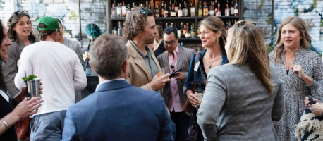 A group of people, both men and women, are socializing at an outdoor bar. Some are holding drinks, engaged in conversation. The background shows a stocked bar with various bottles.