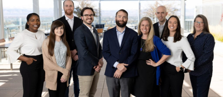 A group of nine people, including four women and five men, stand together in an indoor setting, smiling at the camera. Some people are dressed in professional attire, and the background features large windows.