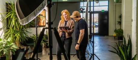 Two people stand in a well-lit studio with plants and lighting equipment, looking at a tablet.