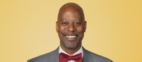 A man in a grey suit and red bow tie smiles in front of a yellow background.