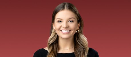 A smiling person with long, light brown hair, wearing a black top and hoop earrings, stands against a red background.