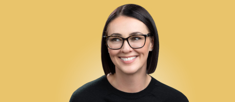 A person with short dark hair and glasses smiles while looking slightly to the side. The background is yellow.