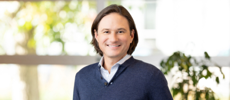 A person with shoulder-length hair and a friendly expression stands indoors in front of a window, wearing a dark blue sweater over a light blue collared shirt.