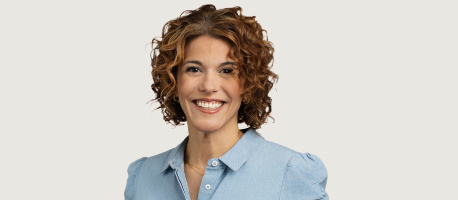 A woman with curly hair smiles at the camera, wearing a light blue button-up shirt against a plain background.