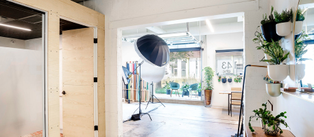 A bright, open office space featuring a photography setup with a large umbrella light, potted plants on shelves, and a glass-walled meeting room. A window and door lead to an outdoor seating area.