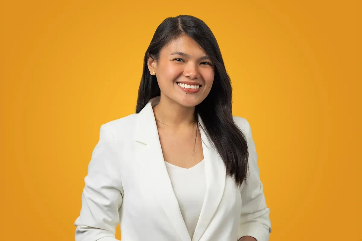 A smiling woman with long dark hair wears a white blazer and stands against a solid orange background.