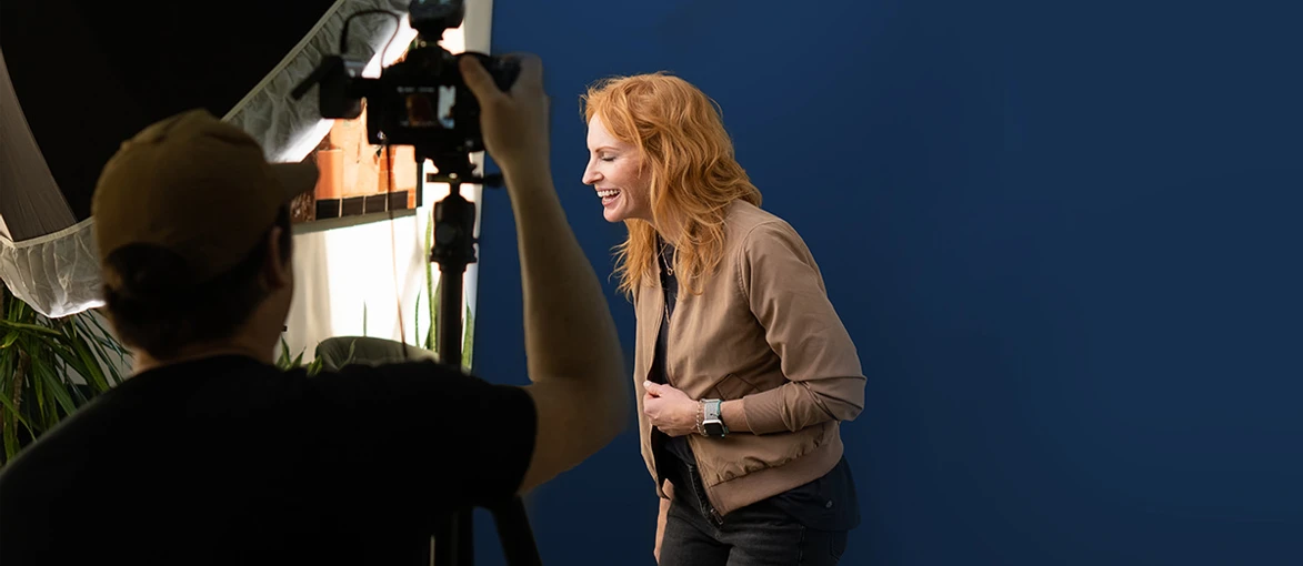 A woman with red hair smiles while posing in front of a blue background as a photographer captures her image.