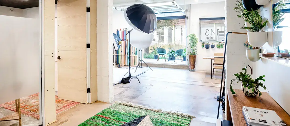 A modern studio with green plants, white walls, colorful rugs, and a photography setup with lights and backdrops. Large windows provide natural light, and shelves line one wall. Door labeled "43" in the background.