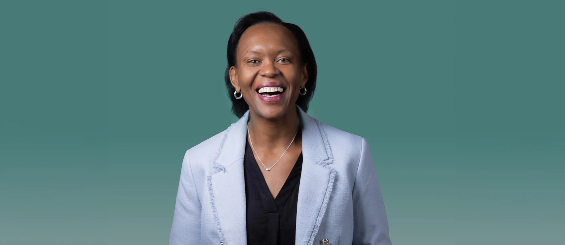 A person with short black hair is smiling while wearing a light gray blazer over a black shirt, set against a teal background.