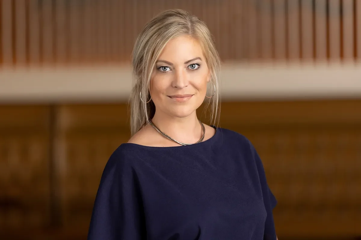 A woman with blonde hair wearing a dark top stands in a room with a wooden backdrop, looking towards the camera.