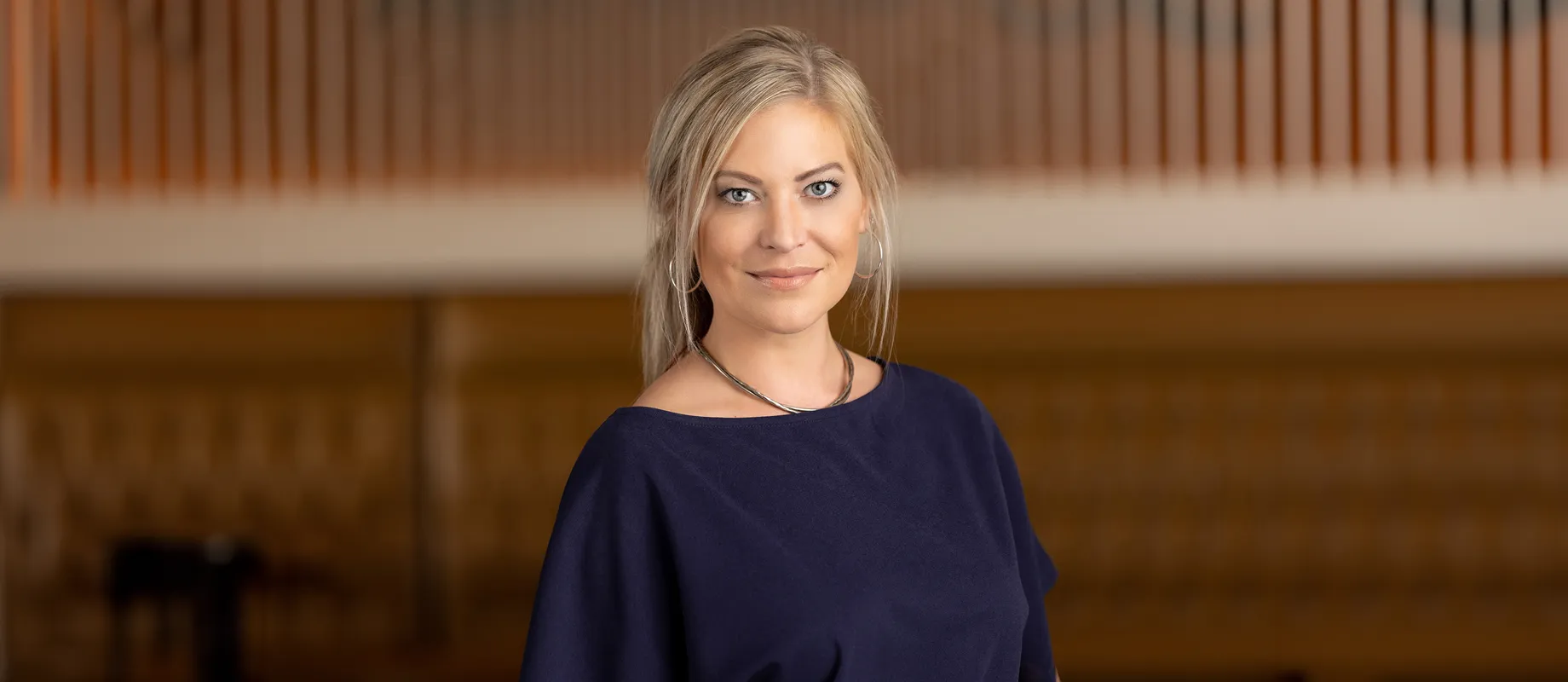 A woman with blonde hair wearing a dark top stands in a room with a wooden backdrop, looking towards the camera.