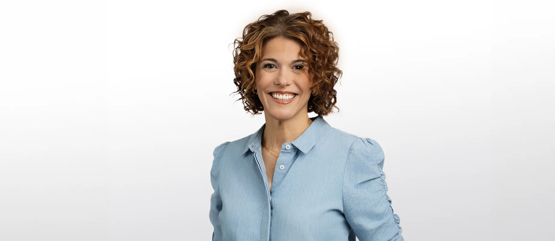 A person with curly brown hair wearing a light blue button-up shirt smiles against a plain white background.