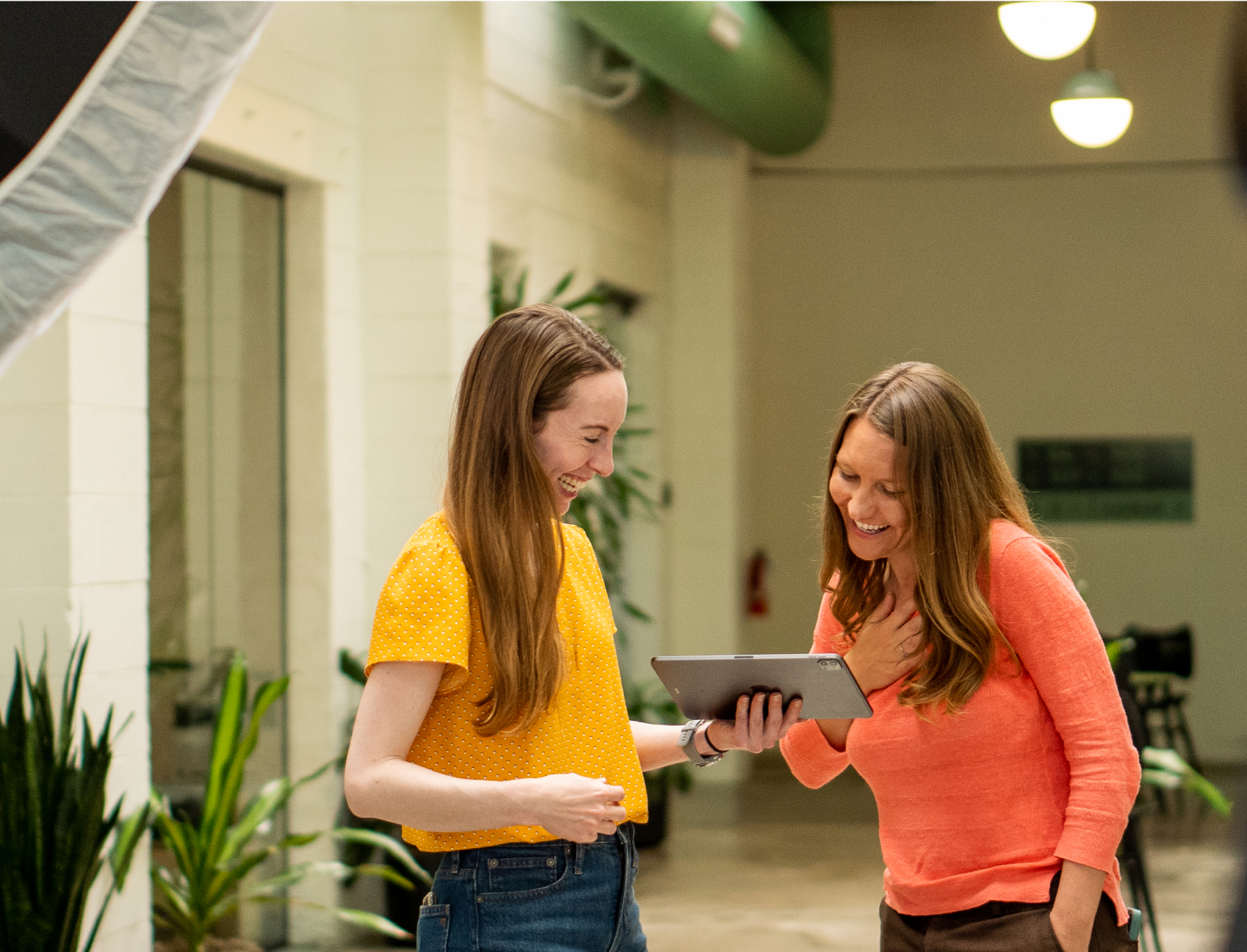 Two women standing indoors in Denver look at a tablet while smiling, surrounded by plants and stylish lighting fixtures—gleefully planning their upcoming event content.