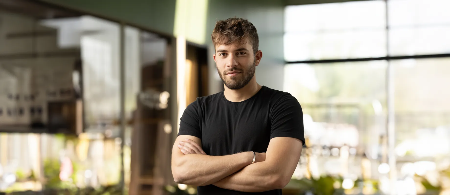 A man with a beard and short hair stands with his arms crossed, wearing a black t-shirt in a well-lit room with glass walls and greenery in the background.