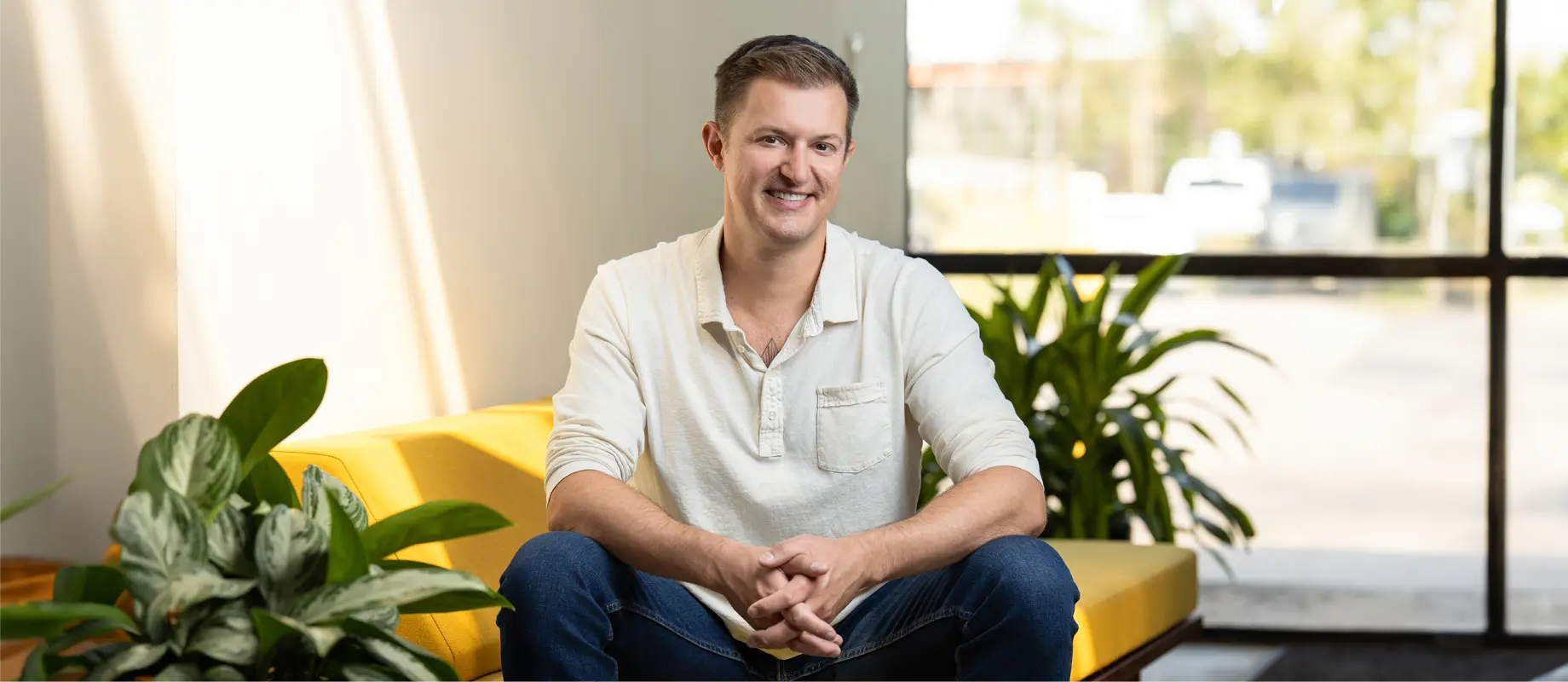 A person wearing a light-colored long-sleeve shirt and blue jeans is sitting on a yellow couch, smiling, with plants on either side and a window in the background.