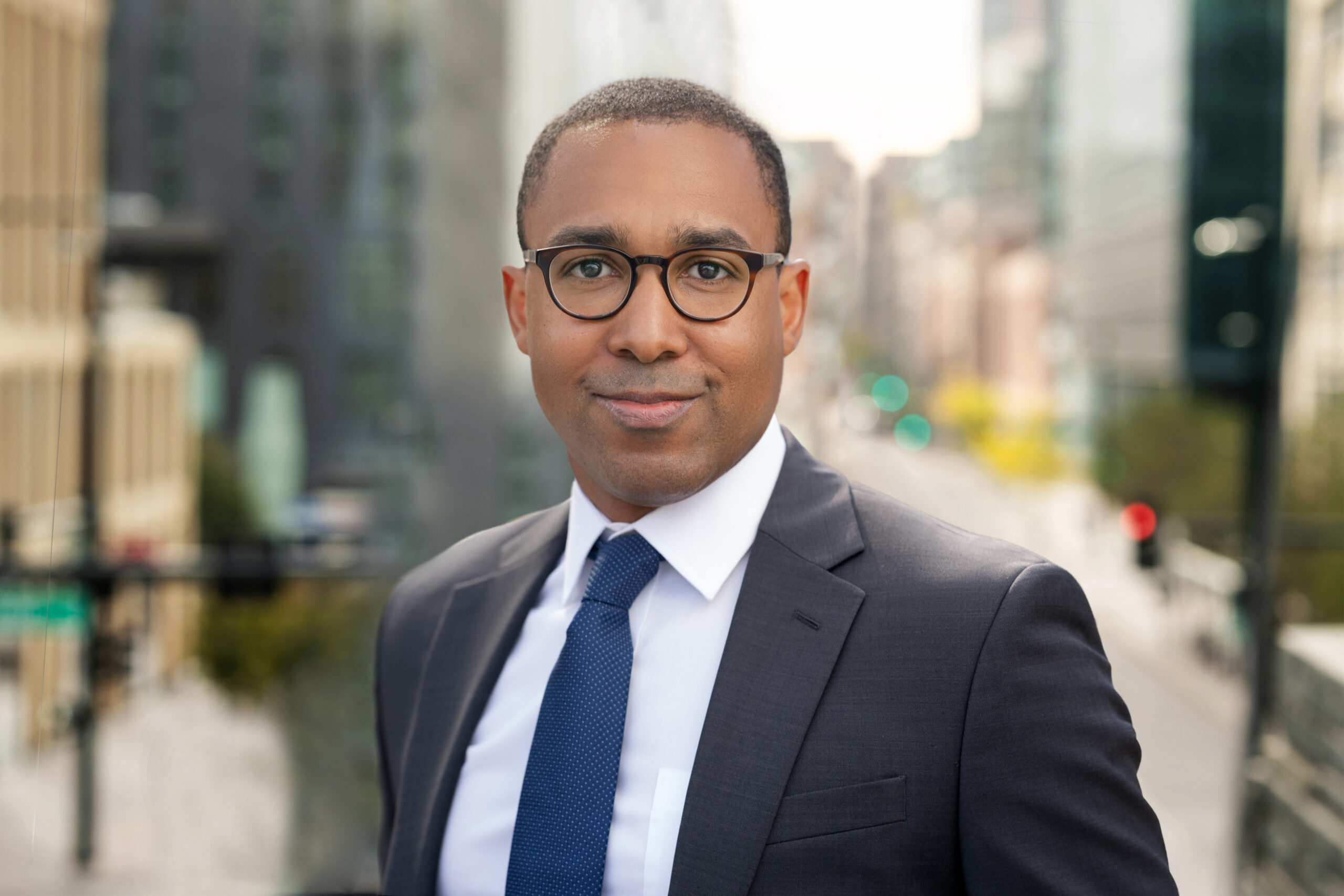 A man in a suit and tie stands outdoors, captured expertly for a headshot photography portfolio, with a blurred urban background.