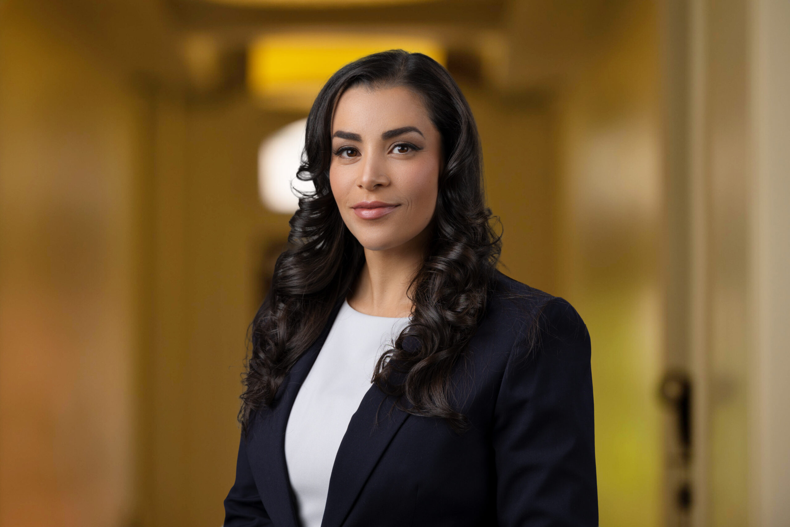 A woman with long dark hair stands in a hallway, clad in a suit, facing the camera with a neutral expression—a perfect headshot for her professional portfolio.