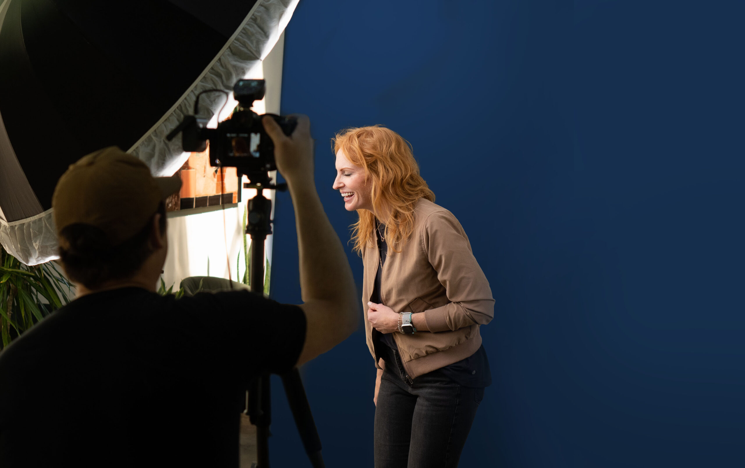 A person with red hair stands and laughs in front of the camera, capturing a dynamic headshot for their portfolio against a blue backdrop.